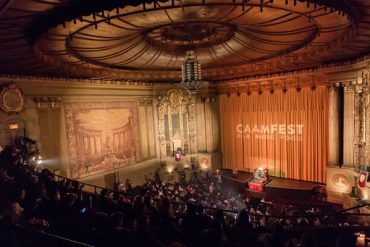 A half-darkened theater, the historic Castro Theater in San Francisco, is filled with audience members waiting for a show to start. Dramatic lighting shines brightly on the curtain, with the words "CAAMFest" on the curtain.
