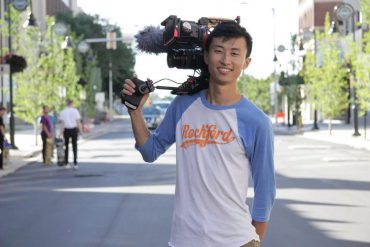 A young Asian American man smiles holding a video camera over his right shoulder.