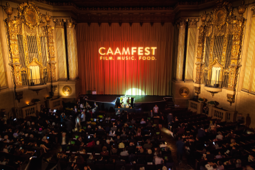 A darkened Castro Theater in San Francisco with the words "CAAMFEST: FILM, MUSIC, FOOD" projected onto the red curtain.