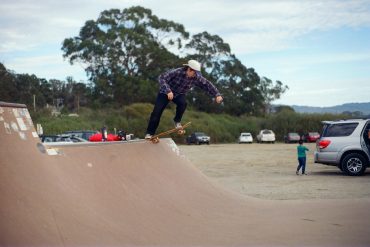 An image of a young Asian American skateboarder getting ready to drop in on a half pipe. The skater is wearing a cap backwards and a flannel shirt.