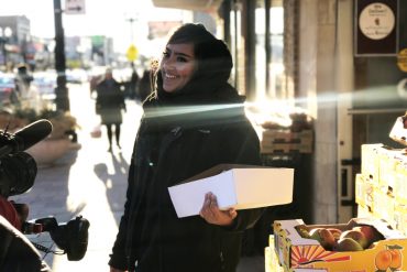 A woman wearing a hijab looks into the distance while holding a box of produce.