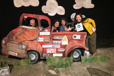 Four people pose joyfully behind an old car in a photo booth. The old car is covered with stickers from nonprofit media arts organizations, including CAAM.