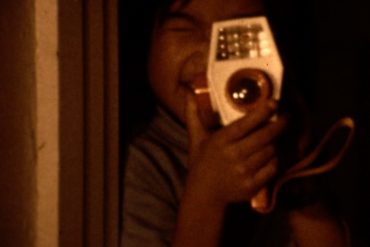 A screenshot of a young Asian American child holding an old video recording device.