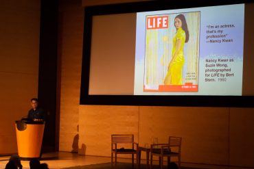 An older Chinese American man stands at the podium with an image of Nancy Kwan on the cover of LIFE magazine projected on a screen.